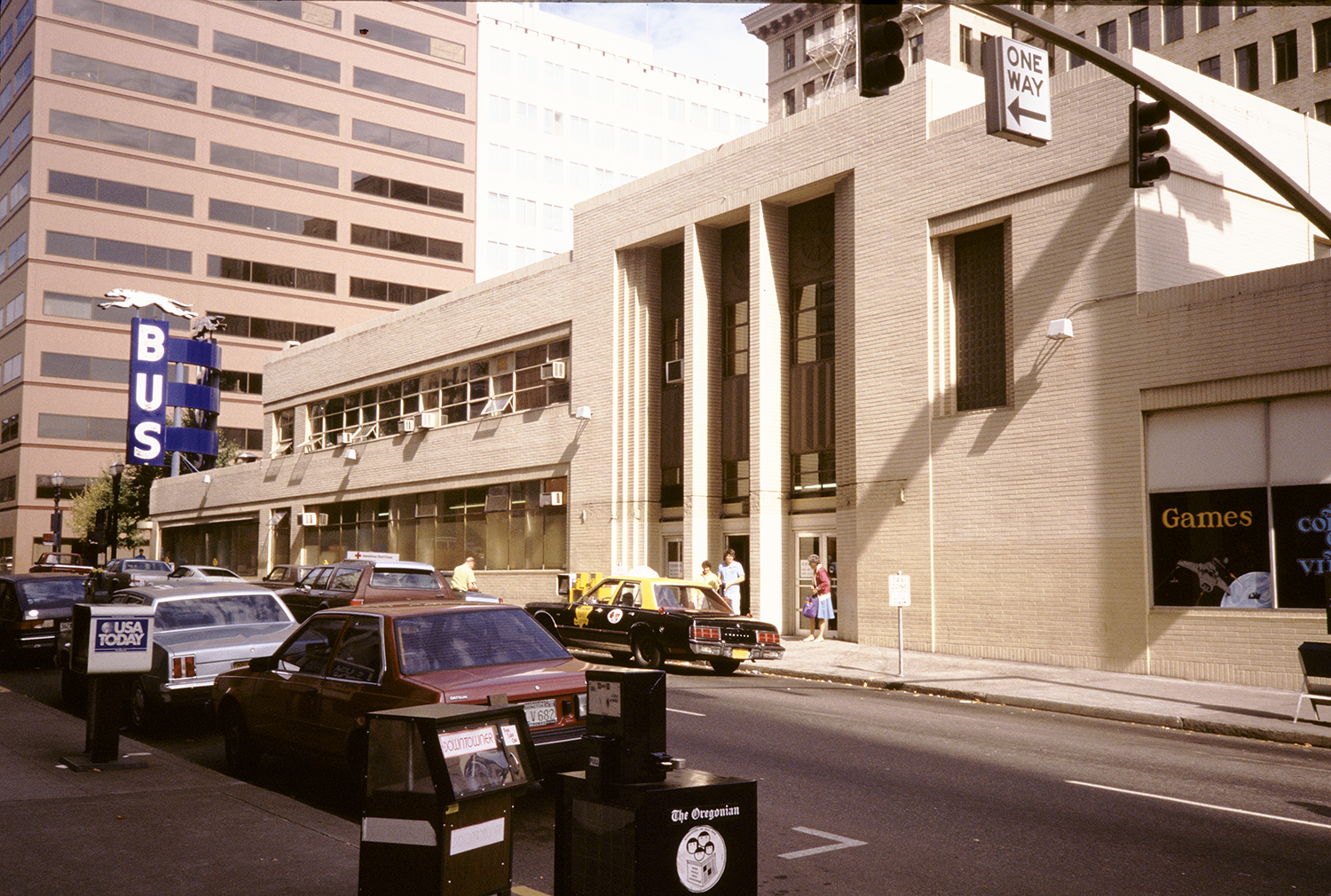 Beige facade, cars, ladies waiting, blue plastic Greyhound sign, window in building says "Games"