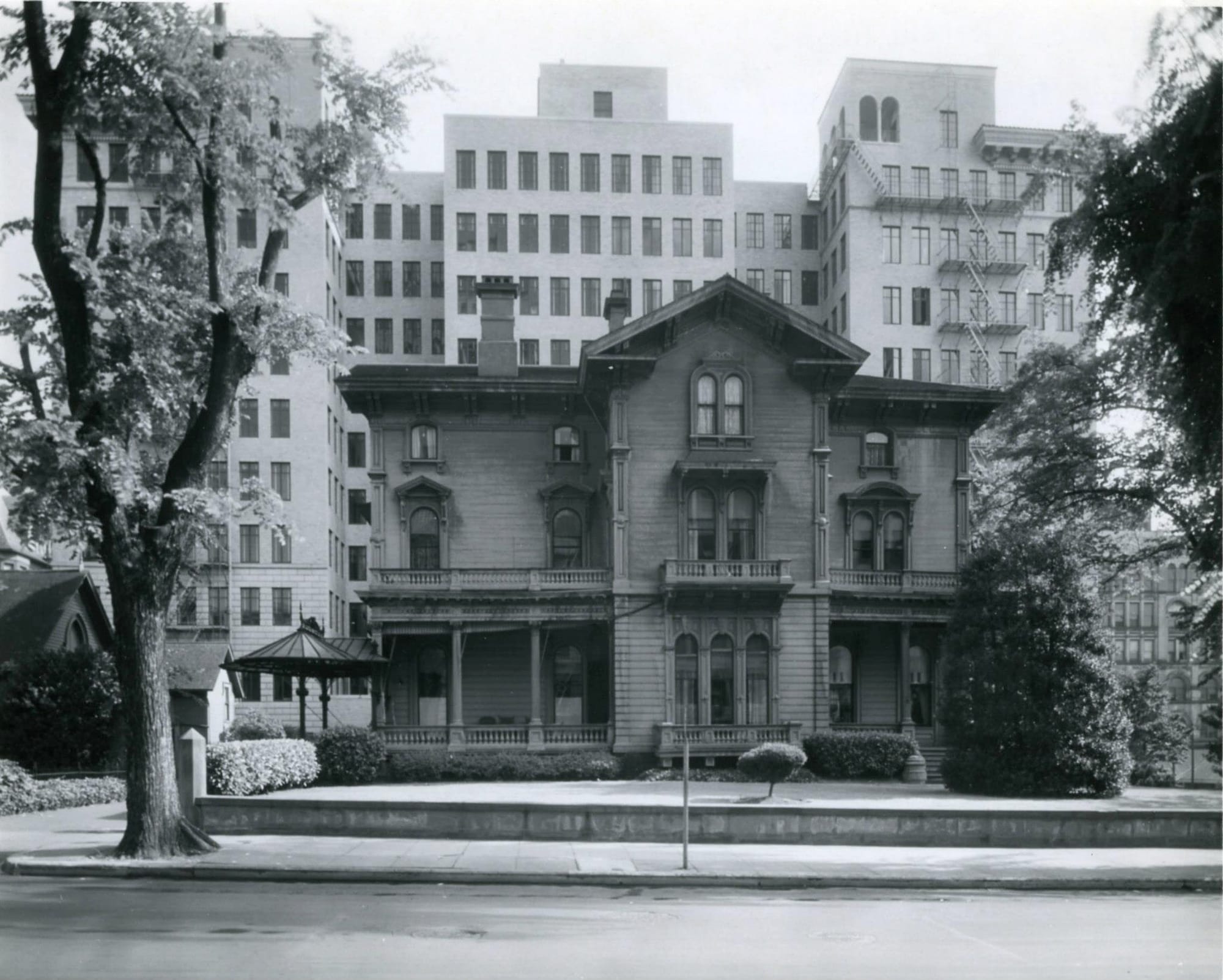 Spooky looking Victorian wooden mansion with the more modern midrise of the Pacific Building in the background. Trees foreground, metal fire escape background.