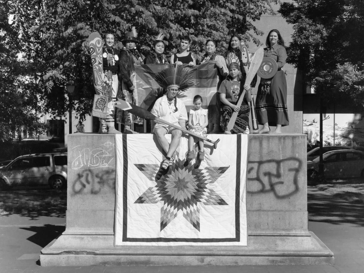 Native Americans sitting on the empty plinth with flags, an oar, and other items.