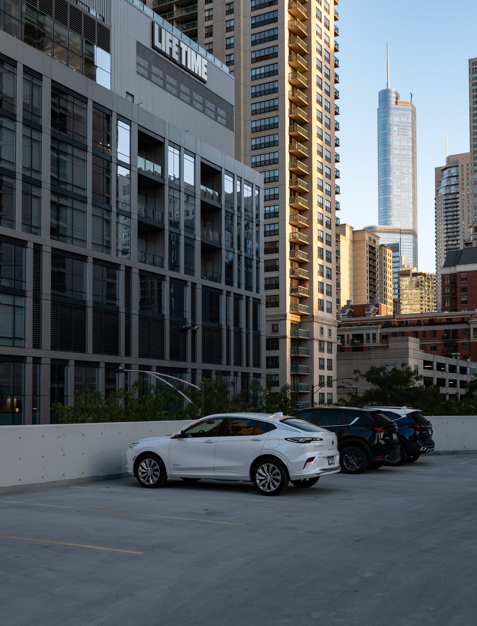 Top floor of a parking garage, white car and black car, Trump Tower in the distance, the One Chicago Square Lifetime Fitness in the foreground.