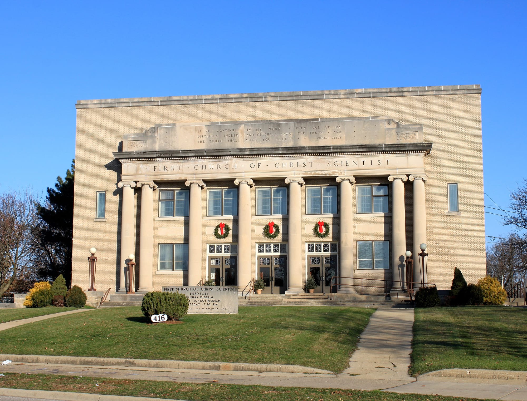 Green lawn with a big bush, church with yellow brick and columns and three Christmas wreaths