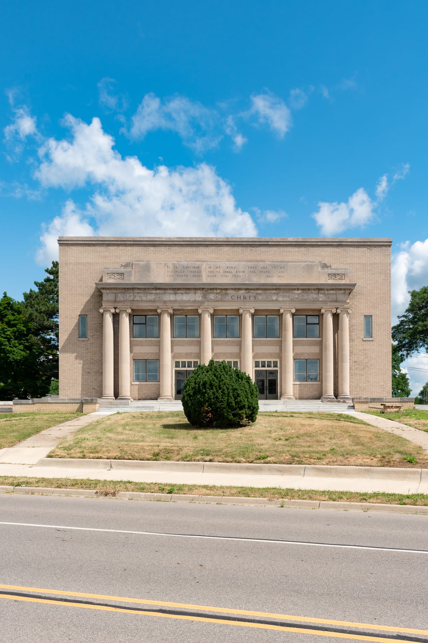 Four lane road, big green bush, boxlike church with eight ionic columns, lettering saying "First Church of Christ, Scientist" has been scratched off